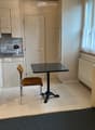 Compact kitchen with light wood cabinets, granite countertop, microwave, sink, small dining table with chair, and vase on counter.
