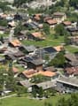Aerial view of a small village with numerous houses, green lawns, and scattered trees. The houses have varied roof styles and colors, and the area is surrounded by mountains.