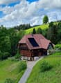 A two-story wooden house with a red-tiled roof, surrounded by lush greenery and hills.