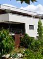 Modern two-story building with white facade, large windows, and a balcony. The surrounding area includes a garden with various plants and a wooden structure, likely part of a residential complex.