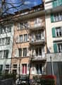 Multi-story residential building with balconies, green shutters, and a tree in front.