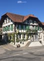 Traditional Swiss building with a red-tiled roof, green shutters, and a balcony adorned with plants. A Swiss flag is displayed on a pole, and a sign reading "Bern" is visible on the left side.