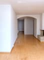 Empty living space with light wooden flooring, white walls, and a fireplace with a tiled surround. A small kitchen area is visible through an open doorway.
