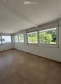 Empty room with tiled floor, white walls, and large windows offering a view of greenery outside.