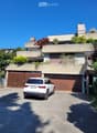 Modern two-story house with a flat roof, large wooden garage doors, and a white SUV parked in front. The house features a raised ground floor and is surrounded by greenery and plants.