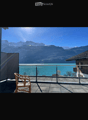 Balcony with a view of a lake and mountains, featuring a single wicker chair and a metal railing.