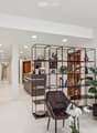 Modern, minimalist waiting area with white walls, a grey sofa, a dark brown leather chair, a large potted plant, a metal shelving unit with books and decorative items, and a reception desk in the background.
