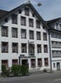 A three-story white building with red-framed windows and decorative trim, featuring a sign reading "Gasthof zur Krone" and a small outdoor seating area with tables and benches.