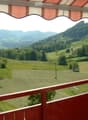 Balcony with red wooden railing, overlooking a scenic valley with green fields, scattered trees, and distant mountains.