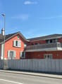 Two-story red house with white shutters, balcony on the upper floor, surrounded by a concrete fence.