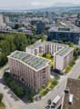 Aerial view of a modern residential complex with solar panels on the rooftops, surrounded by greenery, parking areas, and nearby urban infrastructure including roads, train tracks, and other buildings.
