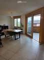 Bright dining area with tiled floor, large wooden dining table, four chairs, a small shelf with decorative items, and a potted plant near the window. A sliding glass door leads to a balcony.