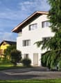 2 story house, white exterior, brown roof, balcony, driveway, car parked, trees, bushes, yellow house in background