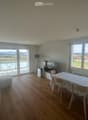Bright living room with wooden flooring, large windows offering a scenic view, a sofa, a dining table with chairs, a television, and a floor lamp.