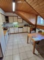 Bright kitchen with white cabinets, wooden ceiling beams, tiled floor, and a dining area with a wooden table and chairs. The kitchen features a refrigerator, dishwasher, and various small appliances on the counter.