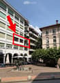 Multi-story apartment building with balconies and green plants, located in a town square with outdoor seating, tables, and a sculpture in the foreground.