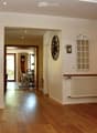 Spacious hallway with wooden flooring, white walls, and a staircase leading upwards. Features a radiator, a wall-mounted clock, and a framed picture. A dining area is visible in the background with a table and chairs.