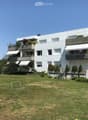 Modern white multi-story building with balconies and greenery, surrounded by a grassy lawn and trees.