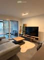 Modern living room with a grey sofa, wooden coffee table, large flat-screen TV, and a guitar leaning against the wall. The room features large windows with blinds and a view of a balcony area.
