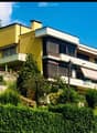A modern two-story house with yellow and white exterior, featuring balconies, large windows, and surrounded by lush greenery and trees.