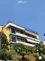Multi-story residential building with balconies, large windows, and surrounding greenery including palm trees and shrubs.