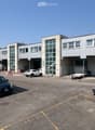 Modern industrial building with large garage doors, multiple windows, and a spacious parking lot with several cars.