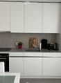 Modern kitchen with white cabinets, grey countertops, and stainless steel appliances. Includes a sink, oven, coffee maker, teapot, and a small potted plant on the counter.