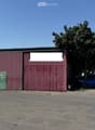 A large industrial-style building with red corrugated metal walls and a green door, situated in a rural area with a tree and a blue car parked nearby.