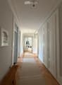 Elegant hallway with herringbone wooden flooring, white walls, and multiple doors leading to different rooms. A framed artwork is displayed on the left wall, and a small rug lies in the center of the hallway.