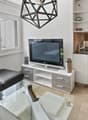 Modern, open-plan living and dining area with a black leather sofa, glass coffee table, flat-screen TV on a white stand, and a compact white kitchen with cabinets, sink, and oven. Large windows offer a city view.