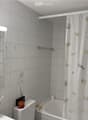A small bathroom with white tiled walls and floor, featuring a bathtub with a shell-patterned shower curtain, a toilet, a mirror with a light fixture above it, and a towel hanging on the wall. Personal care items are placed on the sink counter.
