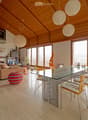 A spacious living room with a high, wooden-beamed ceiling, large windows offering a view, a beige sofa, a colorful ball, a glass dining table with chairs, shelves with books and decorative items, and pendant lamps.