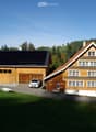 Two-story wooden house with a garage, solar panels on the roof, and a parking area.