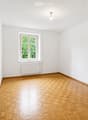 Empty room with wooden parquet flooring, white walls, a single window with white frame, radiator, and a door handle visible on the left side.