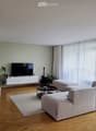 Modern living room with light-colored furniture, a large sectional sofa, ottoman, coffee table, TV, and plants.