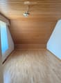 Empty room with light wooden flooring and paneled wooden ceiling, white walls, and a small window with a curtain.