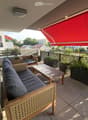 Balcony with a wooden sofa, a small table, a chair, and several potted plants. Features a red awning and a view of the surrounding area.
