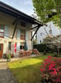 A well-maintained garden area with a wooden dining set, a closed umbrella, a stack of firewood, potted plants, and a flag. The garden is adjacent to a two-story house with light blue shutters and a wooden balcony.
