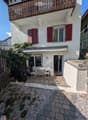 Small outdoor patio area with a table, chairs, and a covered grill, adjacent to a two-story house with red shutters and a balcony.