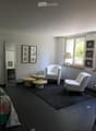 A modern, minimalist living space with white walls, dark flooring, and large windows. Features include a white armchair, small round tables, a floor lamp, and decorative picture frames.