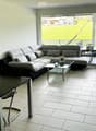 Modern living and dining area with a grey sectional sofa, glass coffee table, black dining table with chairs, and a hanging spherical lamp. Large sliding glass doors open to a view of a green field.