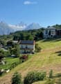 A picturesque village scene with a house featuring a balcony, surrounded by green lawns, trees, and snow-capped mountains in the background.