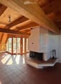Bright, open room with exposed wooden beams, tiled flooring, large windows, and a fireplace. The space is empty, with no furniture or furnishings visible.