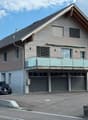 Two-story modern house with wooden siding, large windows, a balcony, and a garage with multiple parked cars.