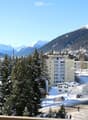Panoramic view of a snowy town with mountains in the background, featuring buildings, trees, and a road.
