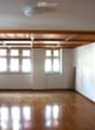 Empty room with wooden flooring, white walls, and large windows allowing natural light. The ceiling features exposed wooden beams, and there is a partially open wooden door leading to another space.