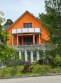 Two-story orange house with a raised ground floor, balcony, and surrounding greenery.