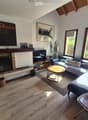 Modern living room with wooden flooring, large windows, fireplace, sofa, coffee table, and decorative items.