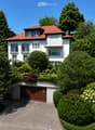 Two-story white house with red-tiled roof, multiple windows, balconies, and a garage surrounded by lush greenery and trees.