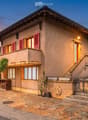 Two-story house with beige facade, red shutters, and a raised ground floor accessed by stairs. Features a stone-paved entrance area, small garden, and a balcony on the upper level.